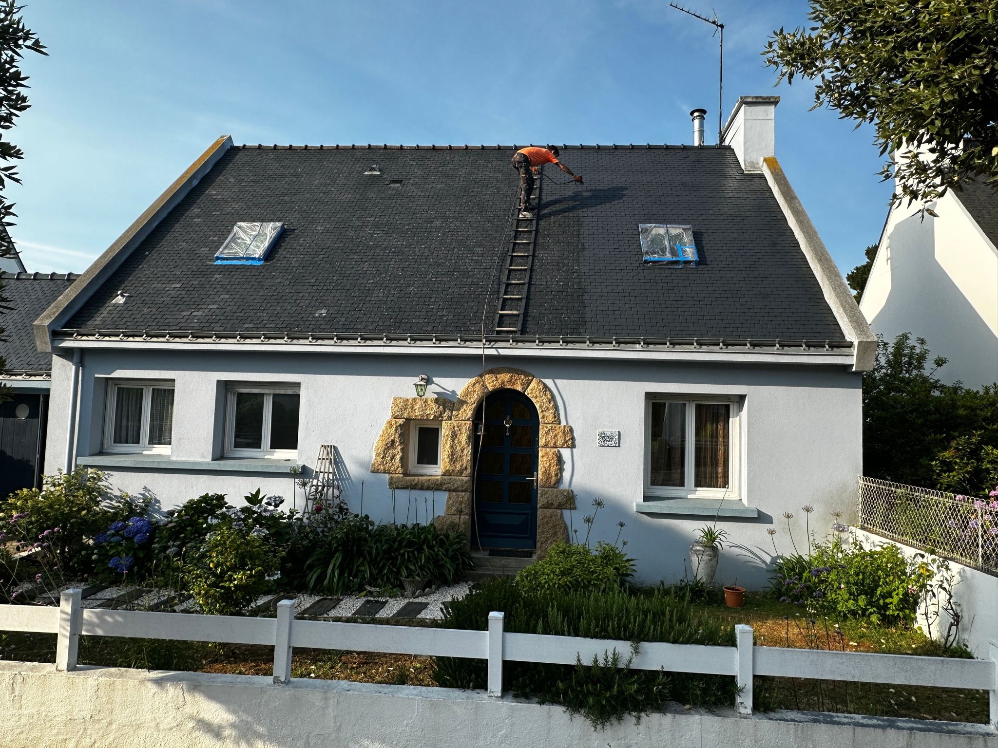 Couvreur traitant une toiture en ardoise à Erdeven depuis une échelle de toit, fenêtres de toit protégées