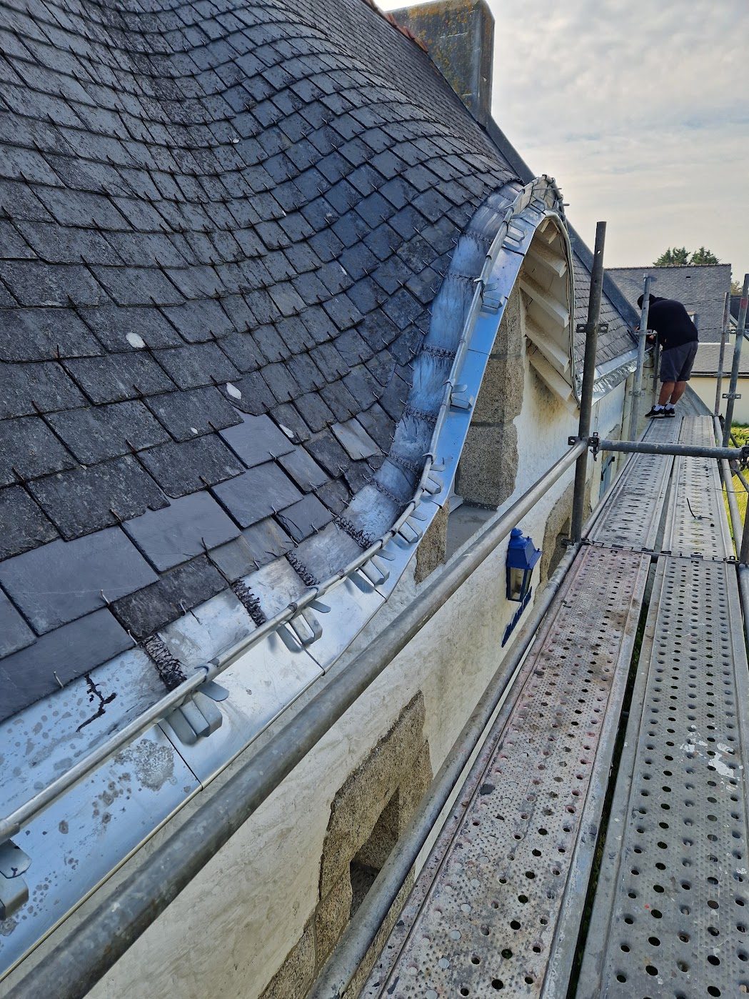 Gouttière en zinc neuve le long d'une toiture en ardoise, avec l'habillage en zinc d'une lucarne cintrée, échafaudage en place